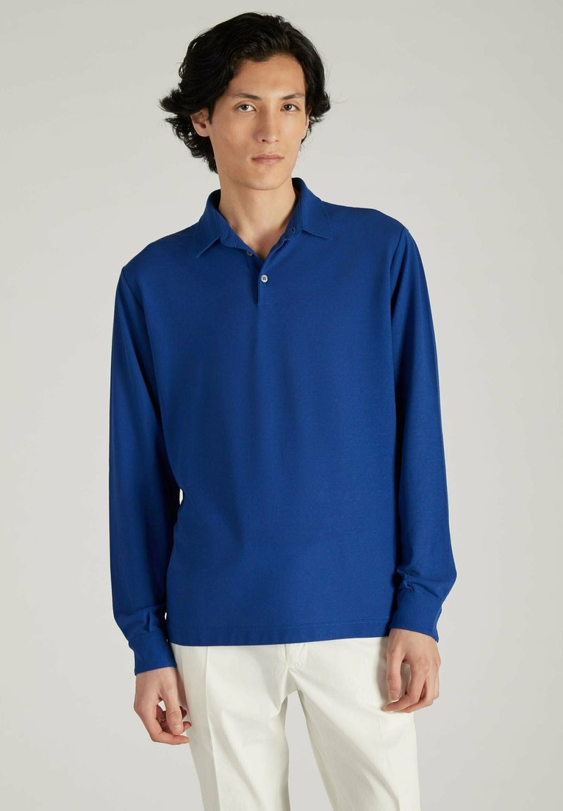 Polo à manches longues de couleur bleu vif, fabriqué en matériau lisse, présentant une patte de boutonnage et un col classique, associé à un pantalon blanc.
