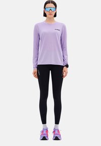 Helle lila Langarmoberteil mit schwarzem Logo, kombiniert mit schwarzen Leggings und bunten Turnschuhen mit pinken und lila Akzenten.