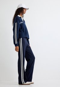 Marineblauer Oversized-Pullover mit weißen Streifen und Logodetail, kombiniert mit passenden marineblauen Jogginghosen mit weißen Seitenstreifen und ausgestellten Beinen.