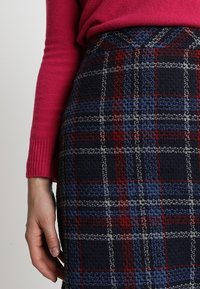 Polvenpituinen, tummansininen ruutuhame, jossa on punaisia ja harmaita yksityiskohtia. Kankaan tekstuuri on ruudullinen. Yhdistetty vaaleanpunaiseen neulepaidat.