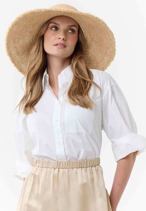 Naine, kellel on pikad lainelised juuksed, kannab laia äärega õlgi kübarat, valget nööpidega särk varrukaid üles keeratud ning beeži elastse vöökohaga seelikut.