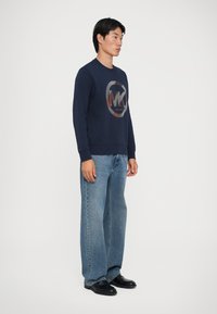 Homem em pé, vestindo um sweatshirt azul-marinho com um logotipo circular, jeans de perna larga em azul claro e sapatos de couro pretos, contra um fundo branco.