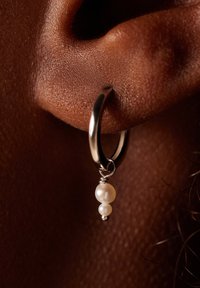 Silberne Creole mit zwei hängenden Perlen; eine große und eine kleine. Der Ohrschmuck wird auf brauner Haut getragen und zeigt eine strukturierte Oberfläche.