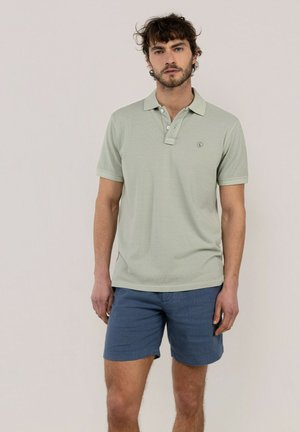 Hombre con camiseta tipo polo de manga corta verde claro y pantalones cortos azules, de pie frente a un fondo beige liso.