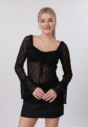 Jeune femme souriante, portant un haut noir en dentelle à manches longues avec des poignets évasés et une mini-jupe noire, les mains jointes à la taille.