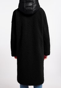 Zwarte, lange jas van gestructureerde stof, met een puffy, gewatteerde capuchon. Het ontwerp heeft een losse pasvorm en een rechte silhoeet.