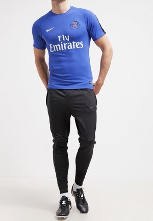 Mężczyzna w niebieskiej koszulce piłkarskiej Paris Saint-Germain, czarnych spodniach dresowych Nike, białych skarpetach i czarnych korkach, stoi ze złożonymi rękami za plecami.