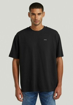 Mann trägt ein schlichtes schwarzes oversized G-Star-T-Shirt und blaue Jeans, steht vor einem neutralen Hintergrund.