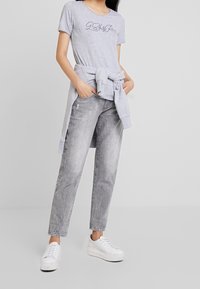 Camiseta gris de manga corta con logo bordado, combinada con jeans desgastados gris claro y zapatillas blancas, atuendo casual.