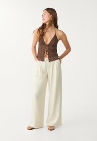 Braunes Haltertop mit geometrischem Muster und vorderem Bindedetail, kombiniert mit crèmefarbenen, weit geschnittenen Hosen. Das Model steht in entspannter Pose.