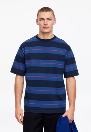 Jonge man die een marineblauw en blauw gestreept T-shirt met dunne rode lijnen draagt, en een blauw kledingstuk in zijn linkerhand vasthoudt tegen een witte achtergrond.