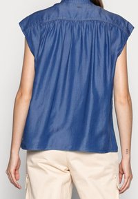 Modrá košile z chambray s límcem na knoflíky, krátkými rukávy typu cap a se shromážděním na zadní straně. Bavlněný materiál s hladkou strukturou.