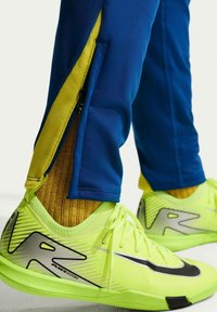 Zapatillas Nike Air Zoom de color amarillo verdoso brillante, combinadas con pantalones azules que tienen cremalleras amarillas y calcetines color mostaza.