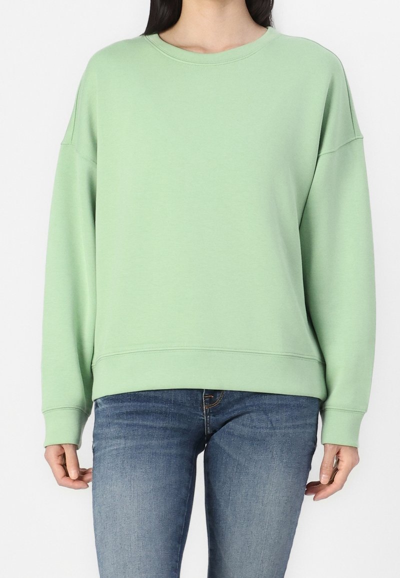Femme portant un sweat-shirt uni vert clair à manches longues associé à un jean bleu, debout sur un fond blanc.