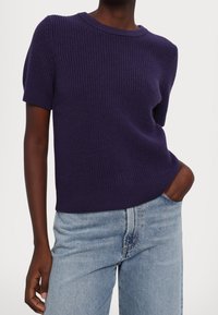 Fialový žebrovaný pletený svetr s krátkými rukávy a kulatým výstřihem, kombinovaný se světle modrými džínami, které mají volnější střih.