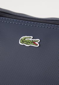 Sac en tissu texturé bleu marine ornée d'un petit logo de reptile vert avec des accents rouges, surligné par une broderie blanche près d'une fermeture éclair.