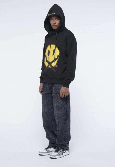 Felpa nera con un grande motivo di faccina sorridente gialla con dettagli neri; abbinata a jeans grigi a gamba larga e sneakers bianco e nere.