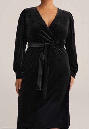 Vrouw draagt een zwarte velvet wikkeljurk met lange mouwen en een ceintuur in de taille, zichtbaar van hals tot halverwege de dij.