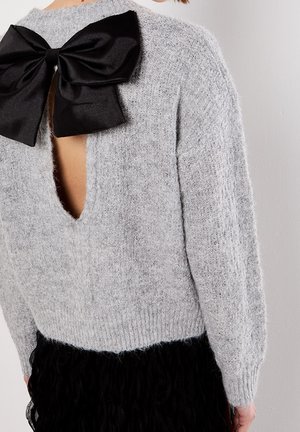 Pull gris duveteux avec dos ouvert et grand nœud noir en satin noué dans le haut du dos, porté avec une jupe noire texturée.