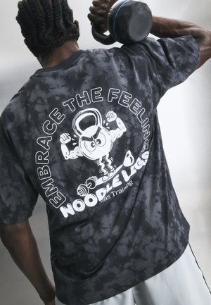 Személy fekete batikos pólót viselve emeli a váll fölé a girját, amelyen izmos rajz és a "Embrace the Feeling Noodles" felirat található.