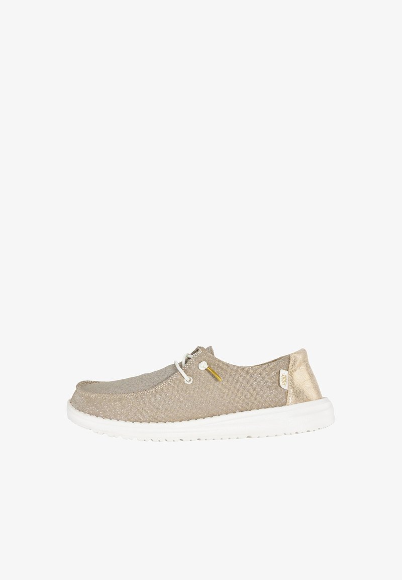 Casual beige instapschoen met witte zool, witte elastische veters, gouden hielaccent en klein stoffen label aan de zijkant.