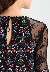 Blouse noire avec un motif floral aux accents rouges, violets et jaunes. Manches en dentelle transparente et un dos à ouverture en forme de goutte avec un lien en ruban.