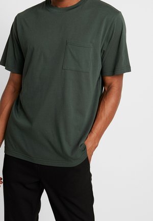 Mand iført mørkegrøn t-shirt med korte ærmer og brystlomme samt sorte bukser, stående foran en ensfarvet lys baggrund.