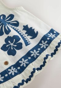 Tissu blanc avec broderie florale et feuillage bleu, un bouton en bois, et un bord froncé avec un galon ondulé bleu.