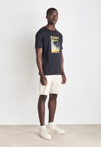 Camiseta negra de algodón con un diseño gráfico de palmeras y colores de atardecer. Combinada con unos pantalones cortos beige claros y zapatillas blancas.