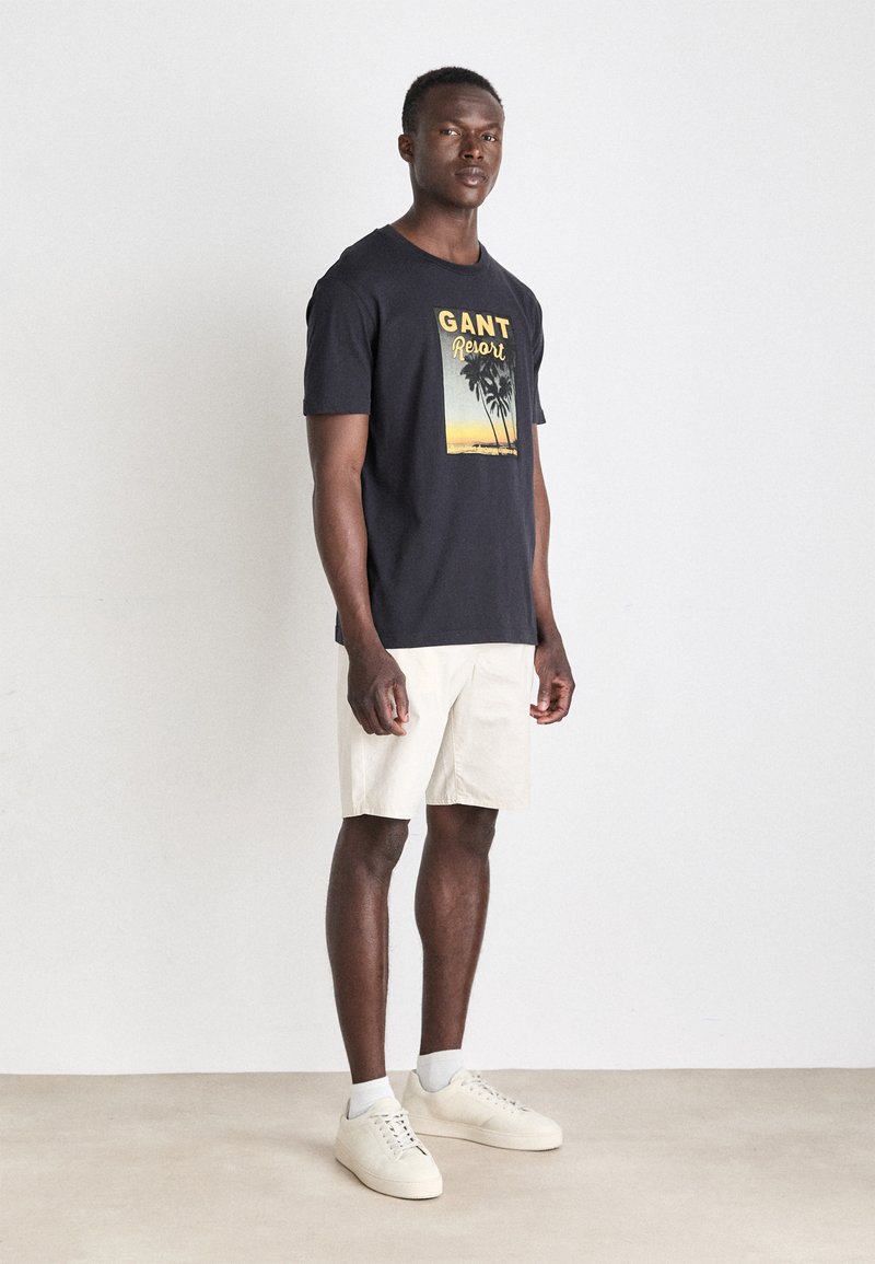 Camiseta negra de algodón con un diseño gráfico de palmeras y colores de atardecer. Combinada con unos pantalones cortos beige claros y zapatillas blancas.