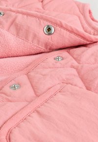 Gilet trapuntato rosa chiaro con morbida fodera in pile, chiusure a snap e fantasie di texture contrastanti. Sono visibili dettagli in hardware argentato.