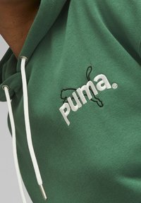 Zielona bluza z kapturem wykonana z bawełny, z białymi sznurkami i haftowanym logo Pumy w kolorze białym i czarnym. Gładka tekstura, swobodny design.