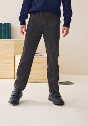 Enfant portant un pantalon gris foncé, des baskets noires à velcro et un pull bleu marine, debout sur un sol clair avec des caisses en bois derrière.