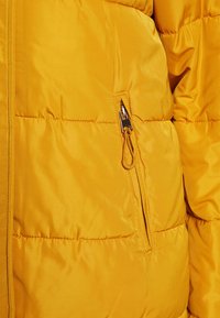 Tissu de doudoune jaune à texture matelassée. Comprend une poche zippée avec une tirette métallique et un détail de cordon élastique.