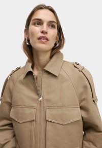 Beige cropped jasje met een stevige kraag, een rits aan de voorkant en twee opgestikte zakken. Het heeft schouderbanden en een gladde, duurzame stoftextuur.