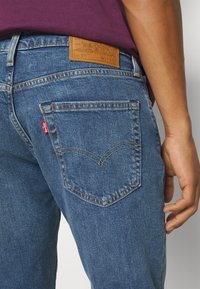Personne portant un jean en denim bleu Levi's avec une poche arrière visible et un patch de marque en cuir à la taille, associé à une chemise violette.