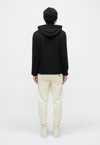 Čierna mikina s kapucňou a predným vreckom, svetlo béžové tepláky a biele tenisky. Mäkká látka, priliehavý strih a malý detail s logom na teplákoch.