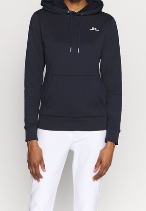 Personne portant un sweat à capuche bleu marine ajusté avec un logo sur la poitrine et un pantalon blanc, debout, les bras détendus le long du corps.