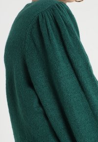 Pull vert en tricot avec une finition texturée, avec des manches volumineuses et des détails froncés à l'épaule.