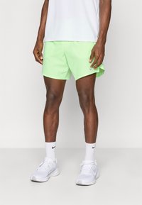 Άνδρας με ανοιχτό πράσινα αθλητικά σορτς, λευκές κάλτσες Nike και λευκά αθλητικά παπούτσια, στέκεται μπροστά σε έναν απλό φόντο.