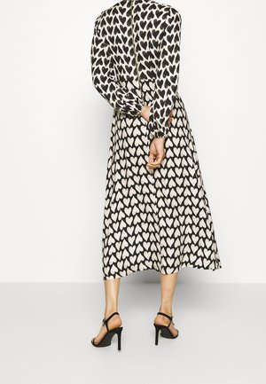 Donna che indossa un vestito midi a maniche lunghe, nero e bianco con motivo a cuori, e sandali neri con tacco alto, in piedi con le mani dietro la schiena.