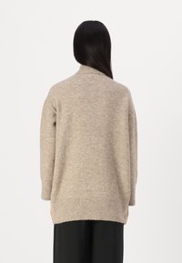 Beige kötött pulóver magas gallérral, bordázott mandzsettákkal és aljjal. Puha tapintású, ejtett vállú és laza fazonú. Nincsenek látható minták vagy fémrészek.