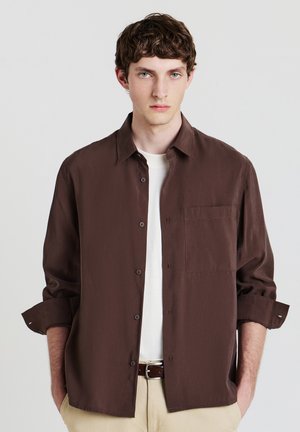 Junger Mann mit kurzen lockigen braunen Haaren, der ein dunkelbraunes Hemd über einem weißen T-Shirt, beigefarbene Hose und einen braunen Gürtel trägt, mit Händen in den Taschen.