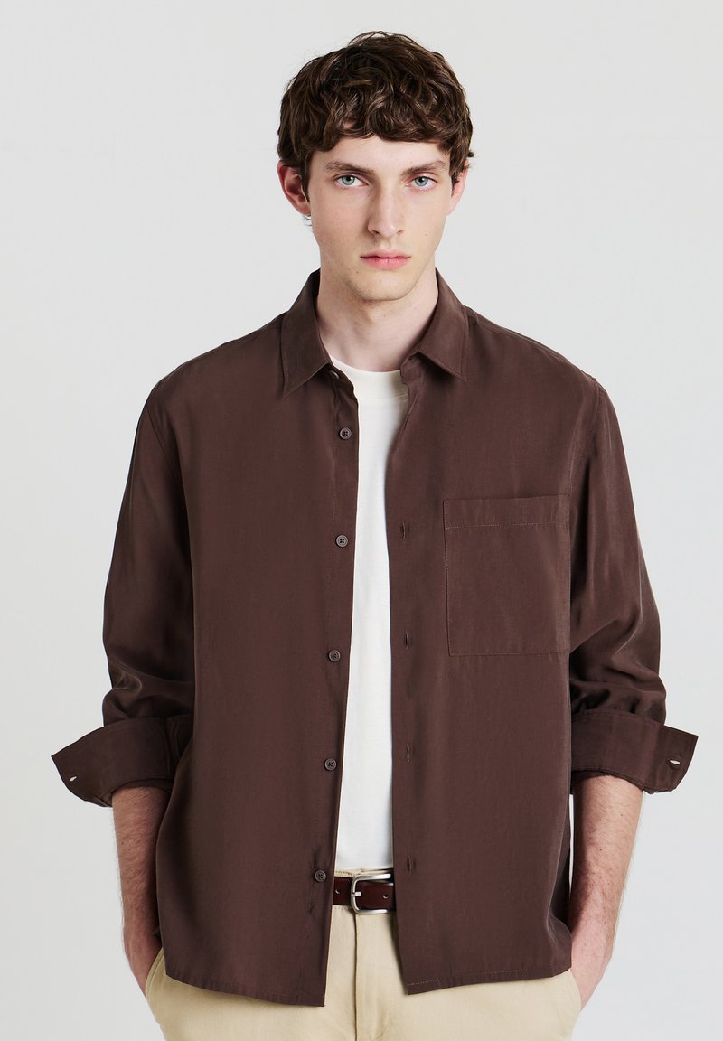 Giovane uomo con i capelli corti e ricci di colore castano, che indossa una camicia a bottoni marrone scuro sopra una maglietta bianca, pantaloni beige e una cintura marrone, con le mani nelle tasche.