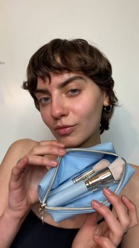 Lyseblå kosmetikkpung med glidelås og sølvdetaljer, som inneholder to børster med myke, lyse børster og slanke, lyseblå håndtak.