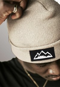 Beige knitted beanie with a ribbed texture and a black rectangular patch featuring a white mountain graphic.