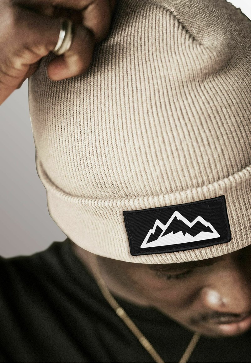 Beige knitted beanie with a ribbed texture and a black rectangular patch featuring a white mountain graphic.