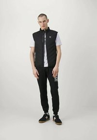 Jovem vestindo um colete acolchoado preto, camisa de manga curta branca, calças de moleton pretas com logotipo e tênis pretos com detalhes brancos.