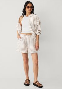 Camisa de lino ligera a rayas con las mangas remangadas, combinada con una camiseta blanca sin mangas y pantalones cortos ajustados del mismo color. Sandalias marrones.