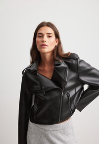 Zwarte cropped leren motorjack met een uitgesneden kraag, meerdere ritszakken en een zachte textuur. Model draagt een grijze trui eronder.